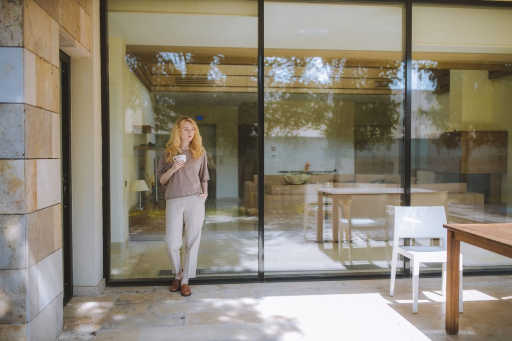 Stylish woman enjoying a coffee while standing by a glass wall in a modern, sunlit room.