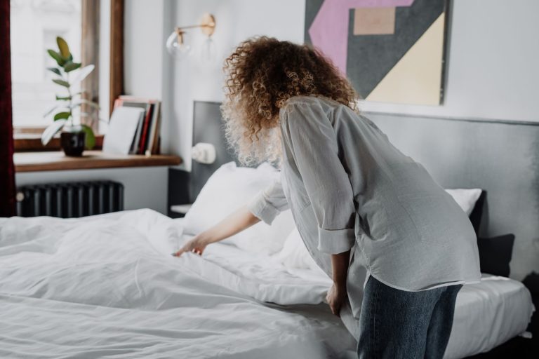 A woman with curly hair making a bed in a modern, cozy bedroom with natural light.