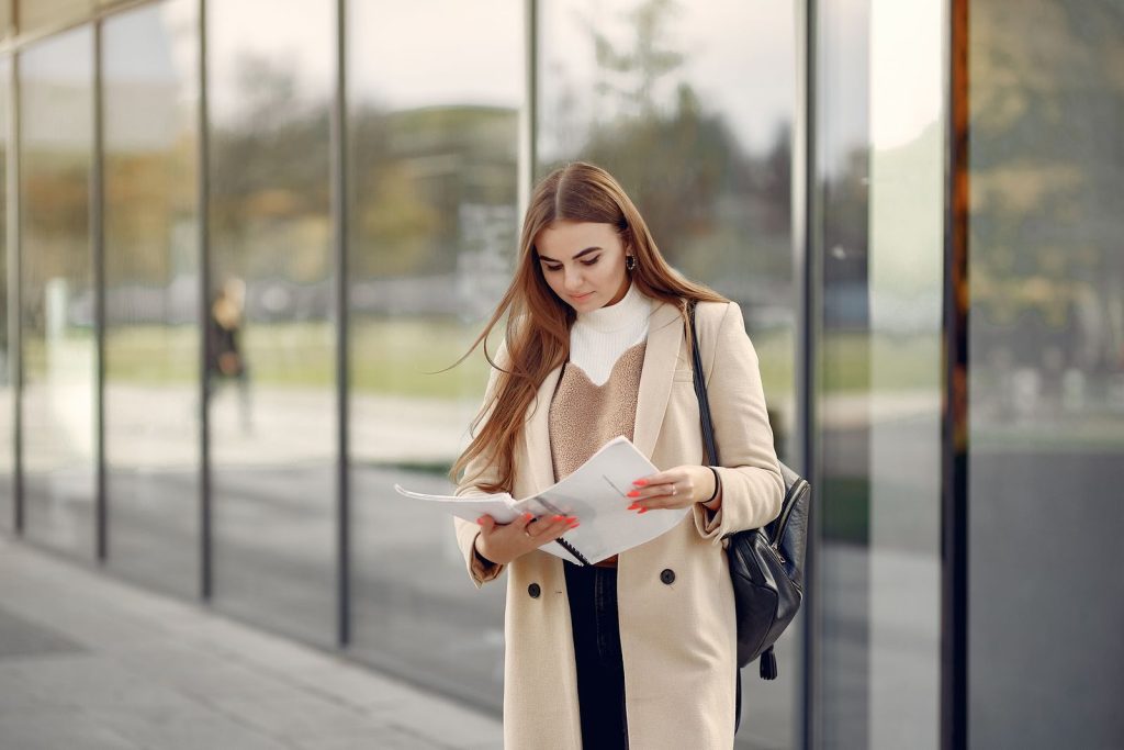 Young female wearing casual clothes standing near glass building and looking at notebook while spending time learning information and thinking