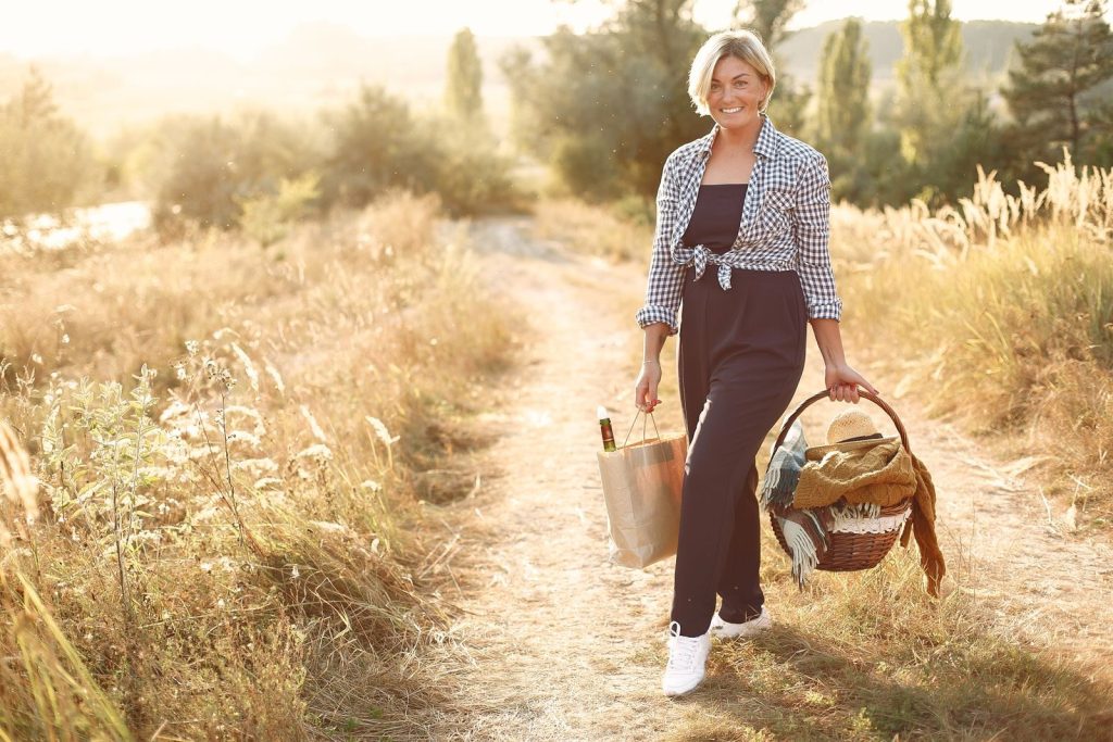 Full body of excited blond woman smiling and looking at camera while walking on path with basket and resting during weekend