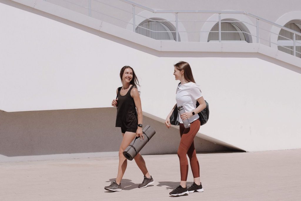 Two women in active wear walking outdoors while smiling and carrying yoga mats.