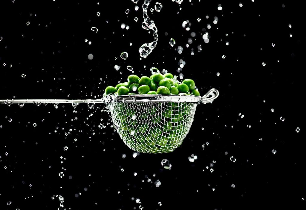 A strainer filled with fresh green peas being rinsed under a splash of water, against a dark background.
