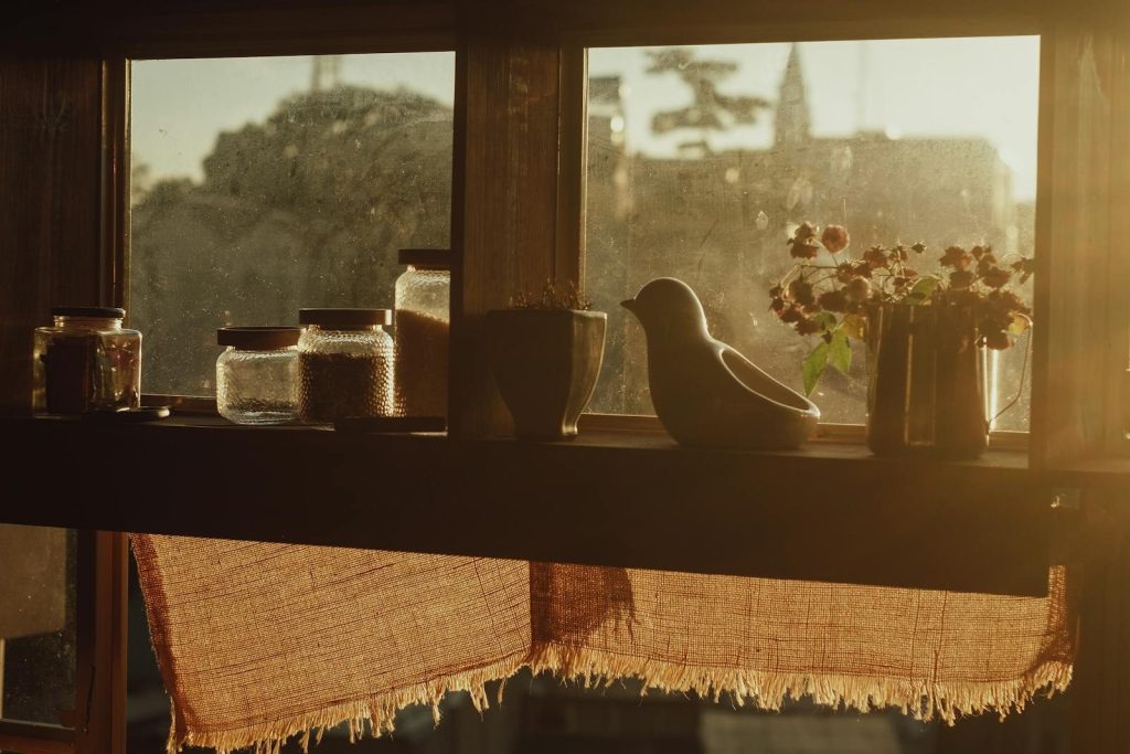 A warm-toned window scene featuring jars, a bird figurine, and flowers, catching sunlight.