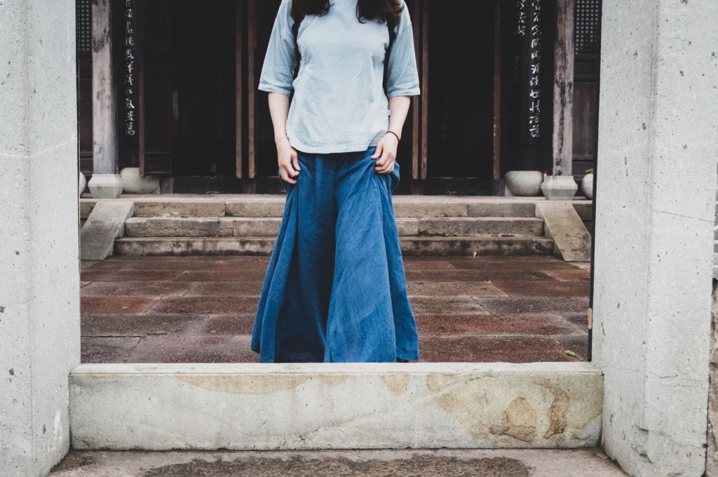 Crop unrecognizable lady in denim wear strolling on shabby tiled pavement near house and concrete fence