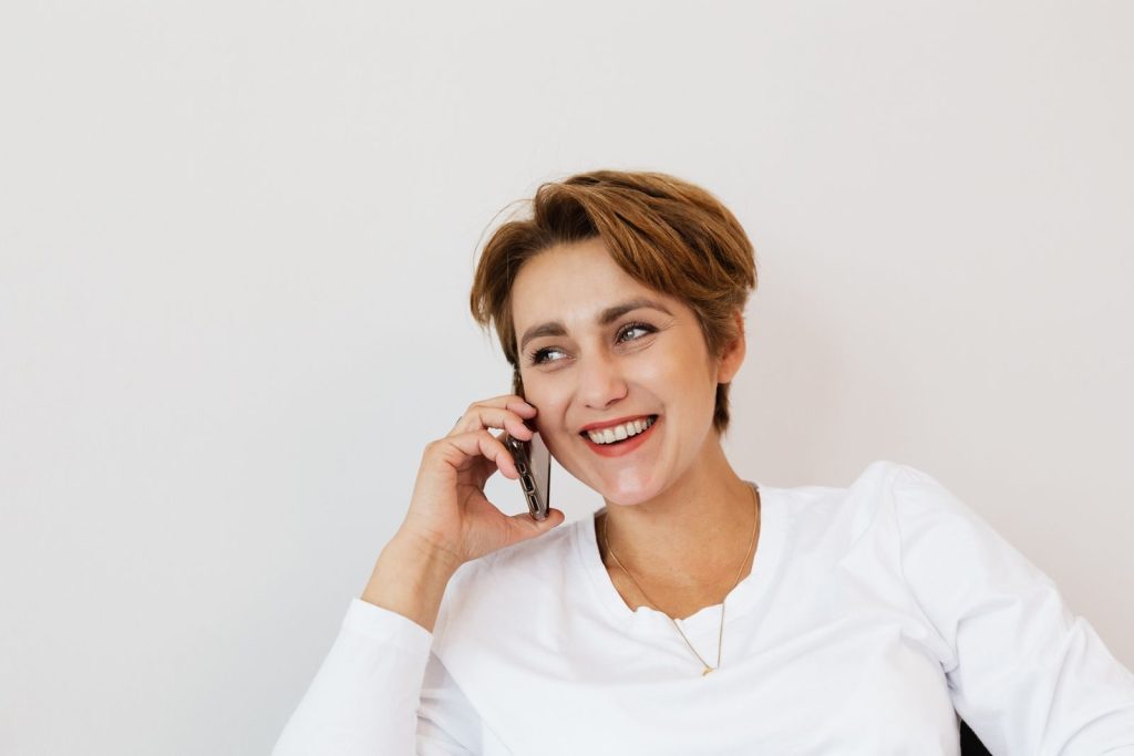 Cheerful female in white shirt laughing while talking on mobile phone and sitting on chair against white wall
