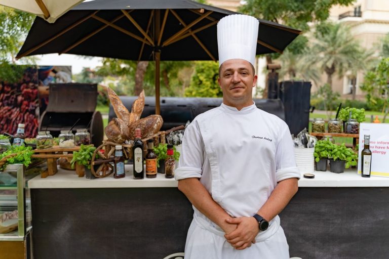 Chef in a white uniform and hat standing in an outdoor restaurant setting in Dubai.