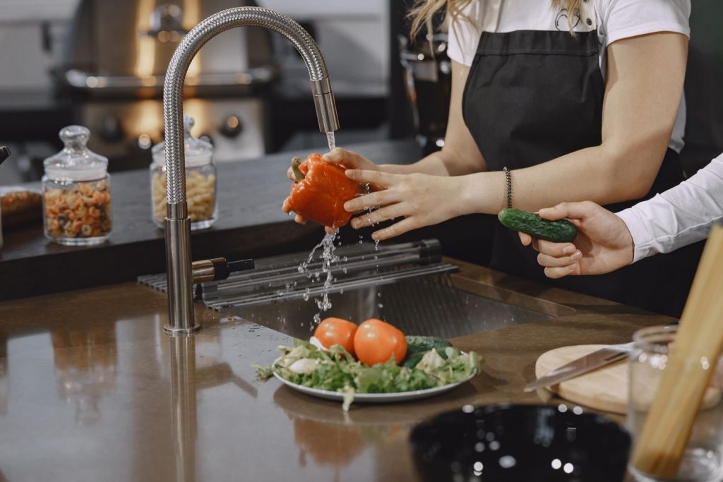 Hands washing a red bell pepper in a modern kitchen sink with fresh vegetables.