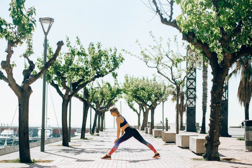 A woman performs stretching exercises on a sunny seaside pathway surrounded by trees.