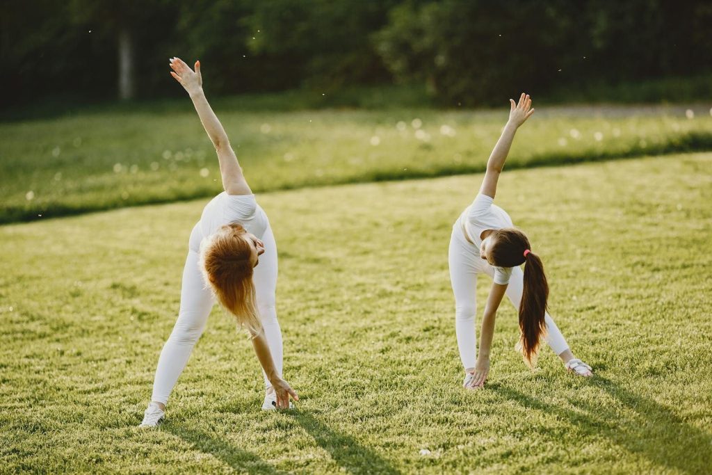 A mother and daughter bond while stretching in a sunny grass field, dressed in white activewear.