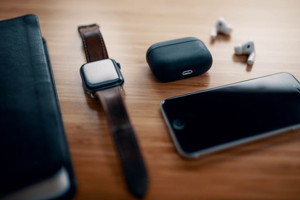 Black smart watch on leather strap and earbuds with charging case and smartphone near black notebook on wooden table