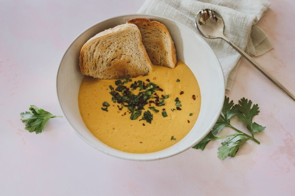 Delicious creamy soup garnished with parsley, served with bread slices on a white surface.