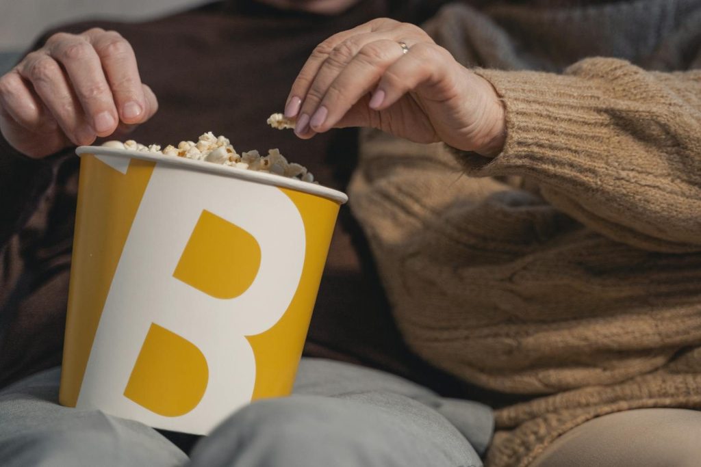 Two adults sharing popcorn from a bucket, wearing cozy sweaters indoors.