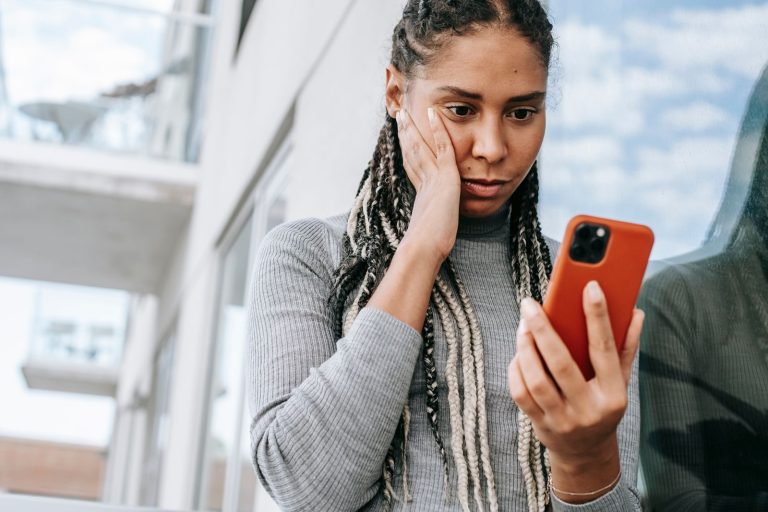 Crop serious African American female with dreadlocks touching cheek in contemplation and browsing mobile phone on street
