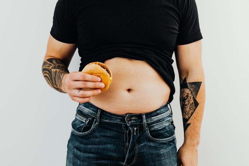 Adult man holding a hamburger, displaying tattoos and abdomen, wearing casual clothing.