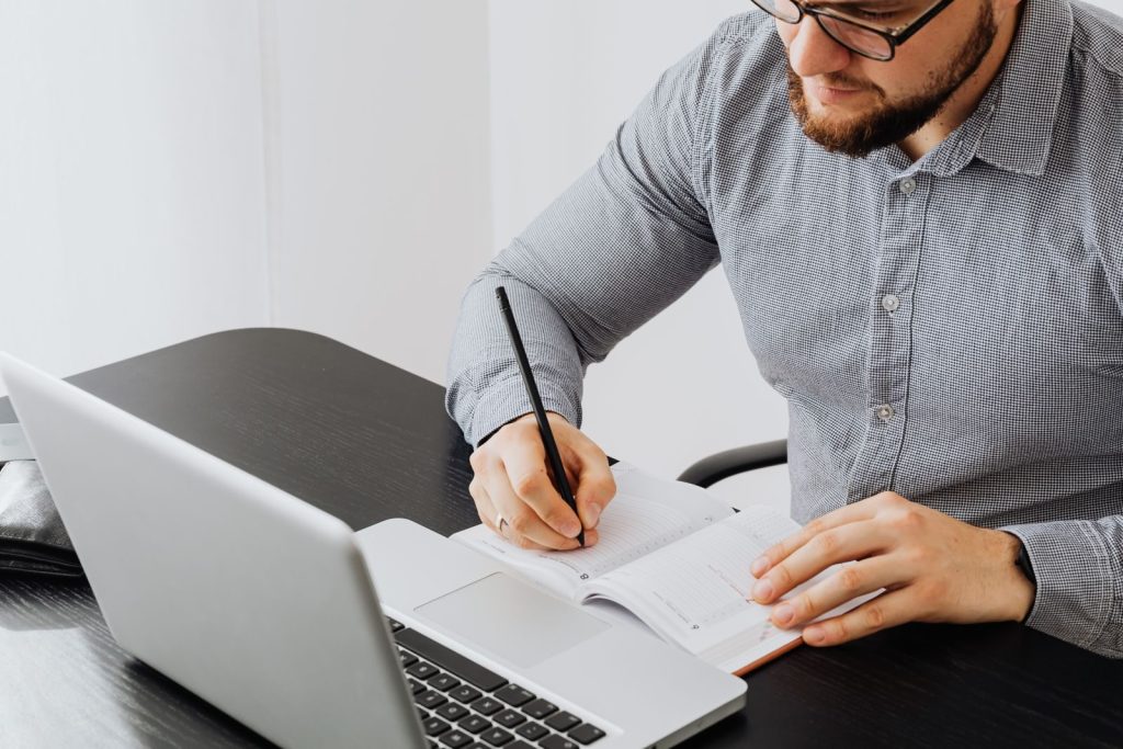 Adult man working with laptop and writing in notebook at a modern indoor office.