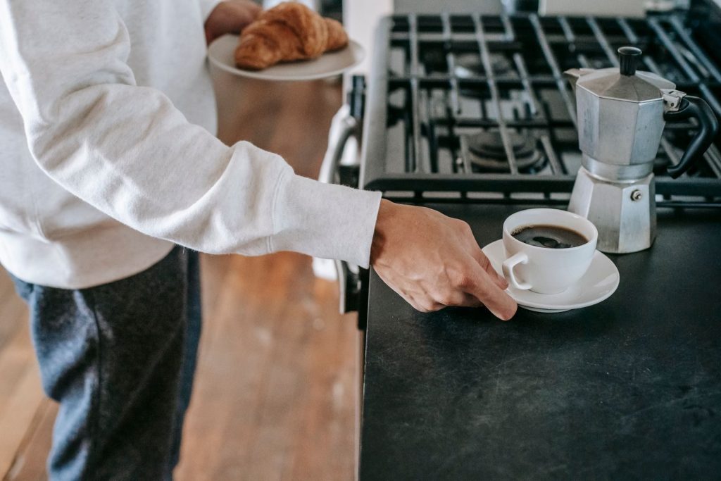 Side view of crop faceless young guy in stylish outfit holding plate with fresh crispy croissant and cup of hot brewed coffee while standing in kitchen during breakfast
