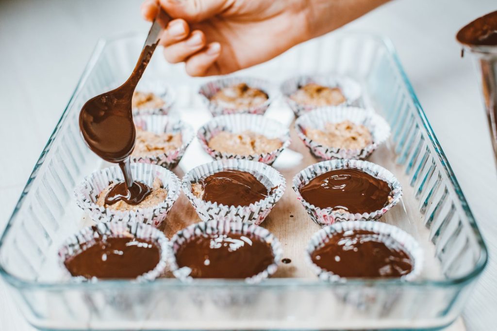 A person delicately pours chocolate over cupcake batter in paper liners, preparing a sweet treat.