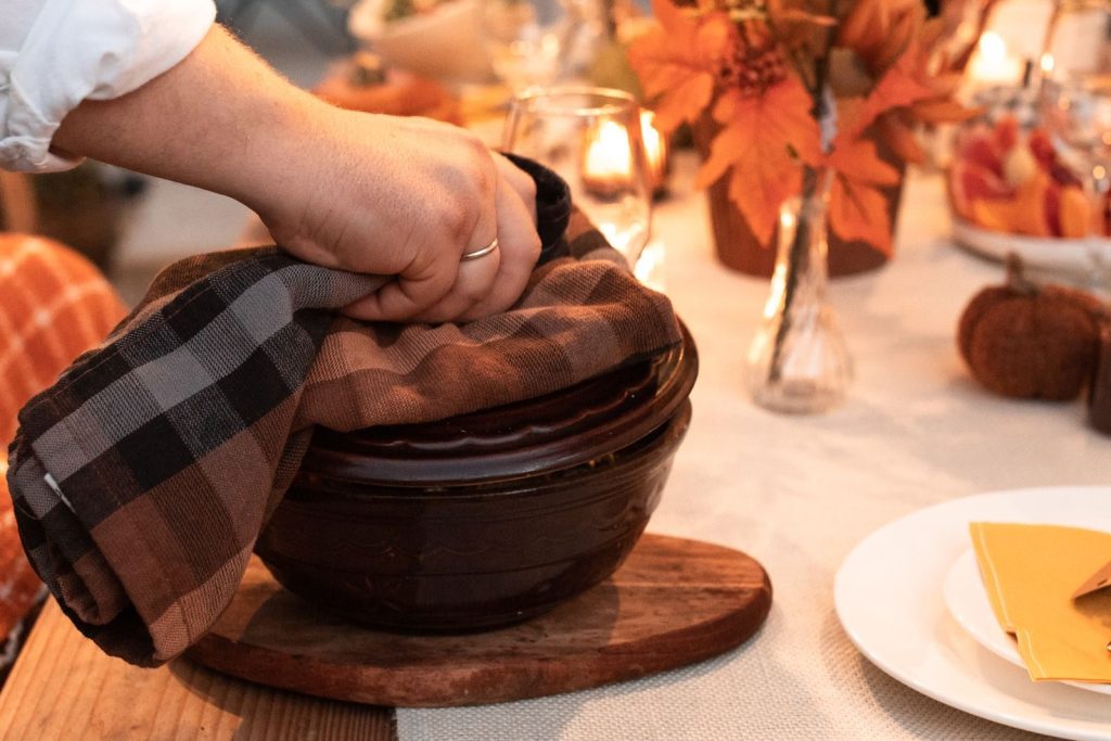 A warm, inviting autumn dinner setting with a hand lifting the lid from a ceramic dish.