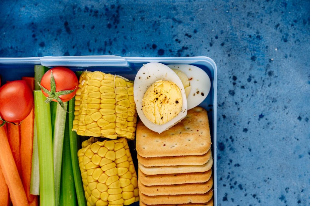 A colorful lunchbox featuring boiled eggs, corn, crackers, and fresh vegetables on a blue background.