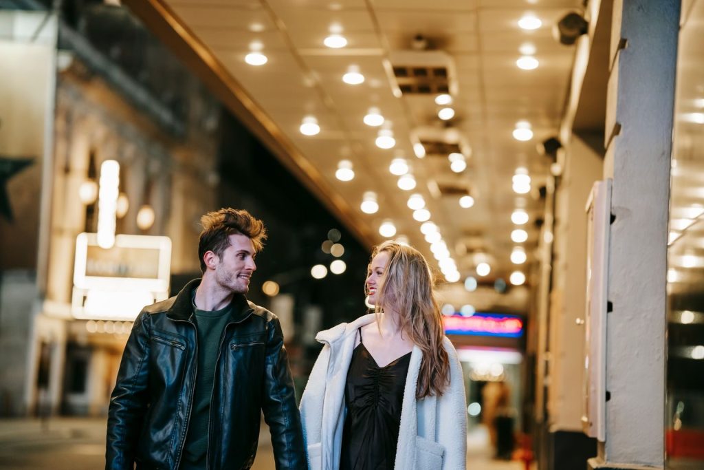 Romantic couple smiling and looking at each other while strolling in hallway of subway station with glowing lights on ceiling on blurred background in evening time