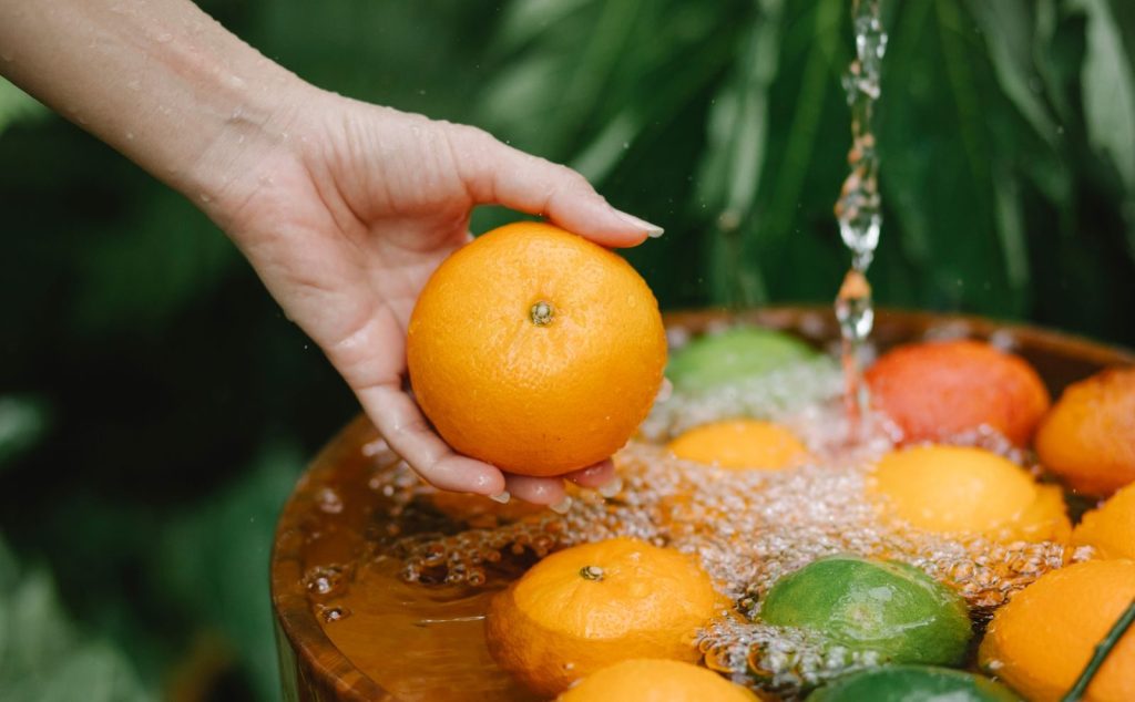 Crop anonymous female washing juicy mandarins under water stream above wooden bowl in lush summer garden