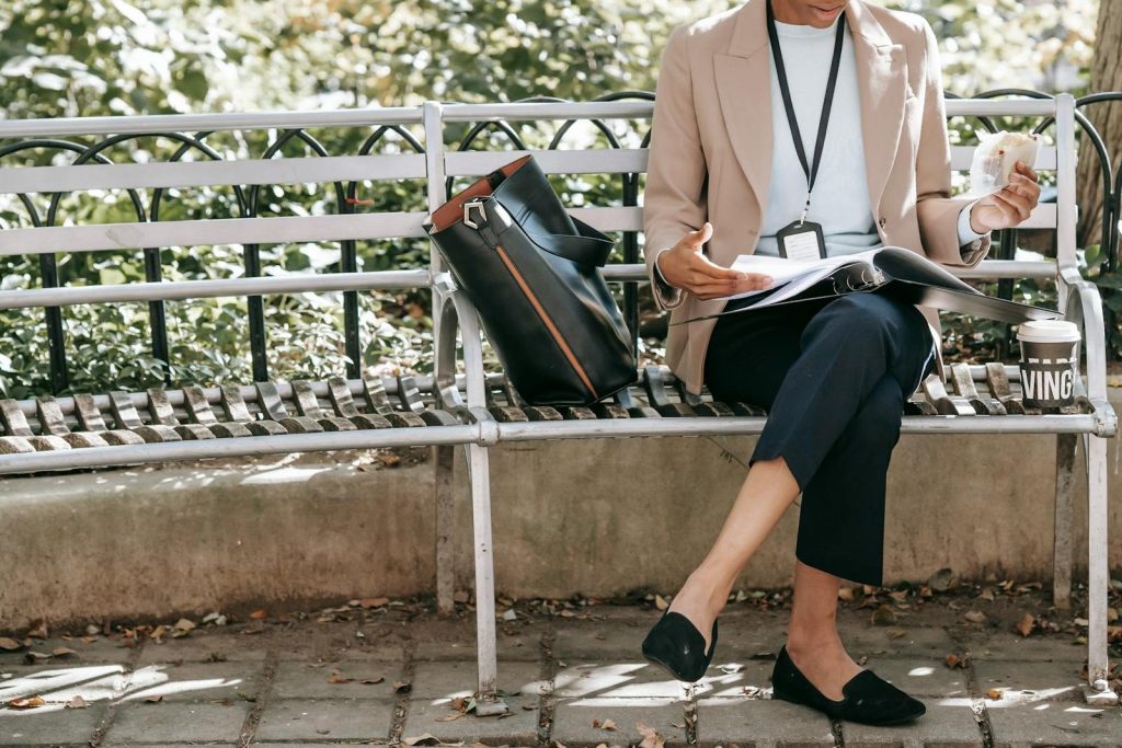 Crop unrecognizable female student reading papers in folder while eating sandwich on bench in park