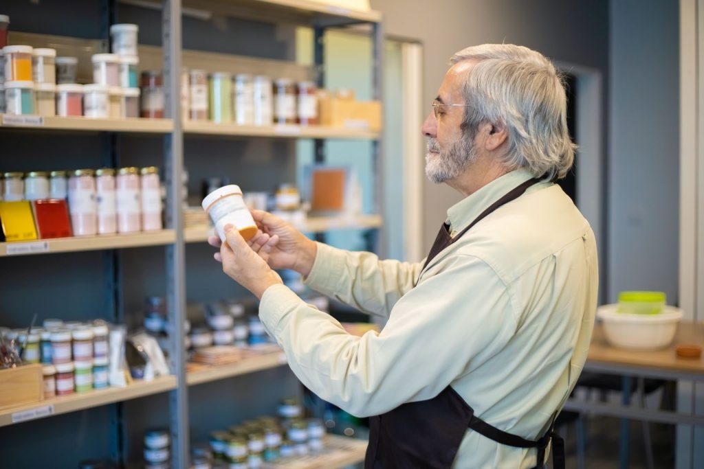 Senior man in apron organizing paint jars on a store shelf indoors.