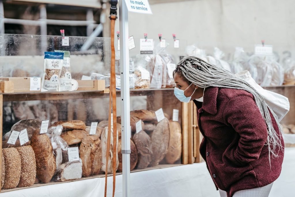 Side view of adult black lady wearing protective mask and warm coat selecting baking goods while standing in street market in daytime