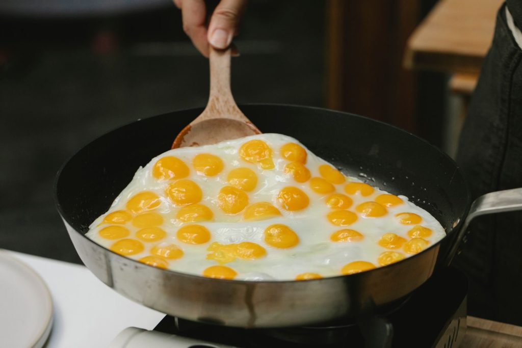 Crop faceless cook with wooden spatula turning quail eggs frying on pan in kitchen