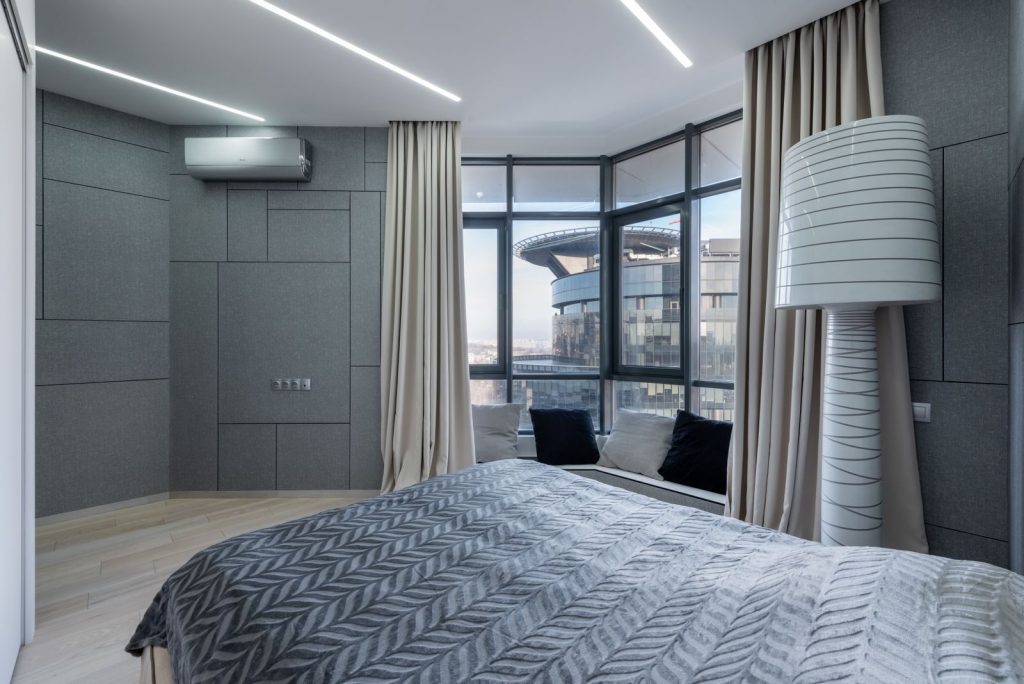 Big bed with blanket placed against gray wall in contemporary bedroom with decorations near window and bright backlight on ceiling