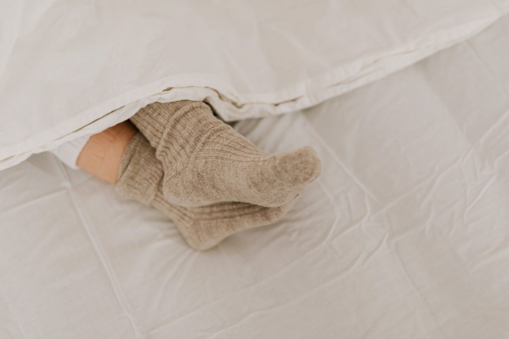 Comfortable feet in woolen socks peek out under a soft white linen blanket on a bed.