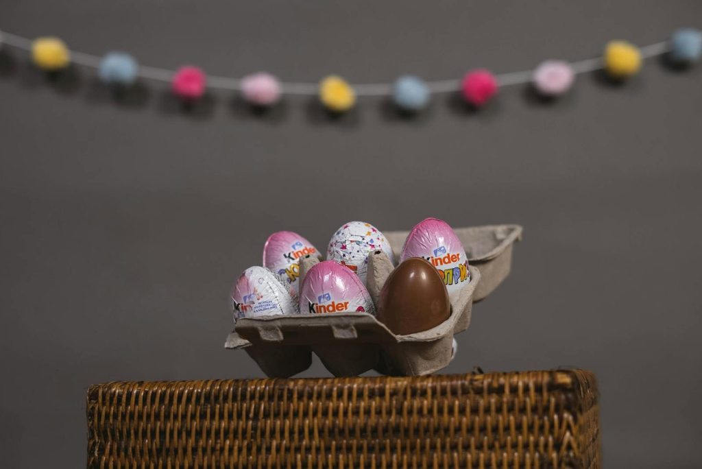 Colorful Kinder eggs displayed in a carton on a woven basket against a gray backdrop.