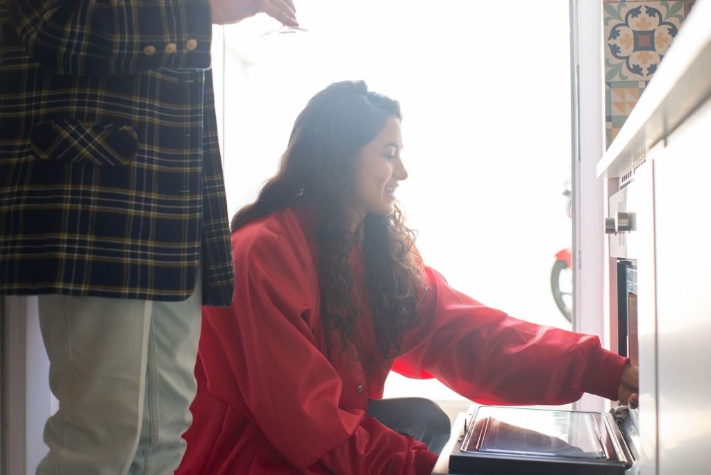 Woman in a red shirt operating an oven indoors, smiling in a bright kitchen environment.