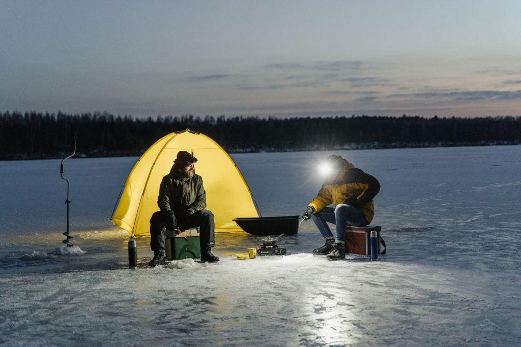 Two adults ice fishing at night near a glowing yellow tent on a frozen lake.
