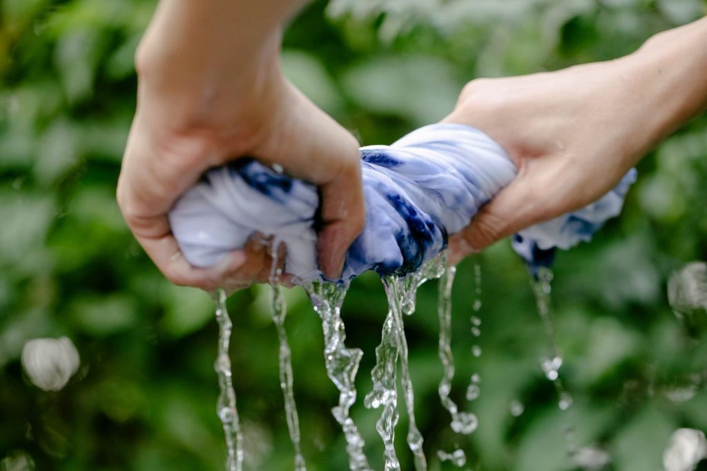 Hands of faceless person with wet dyed fabric in hands with waterdops standing near green plants on blurred background in countryside