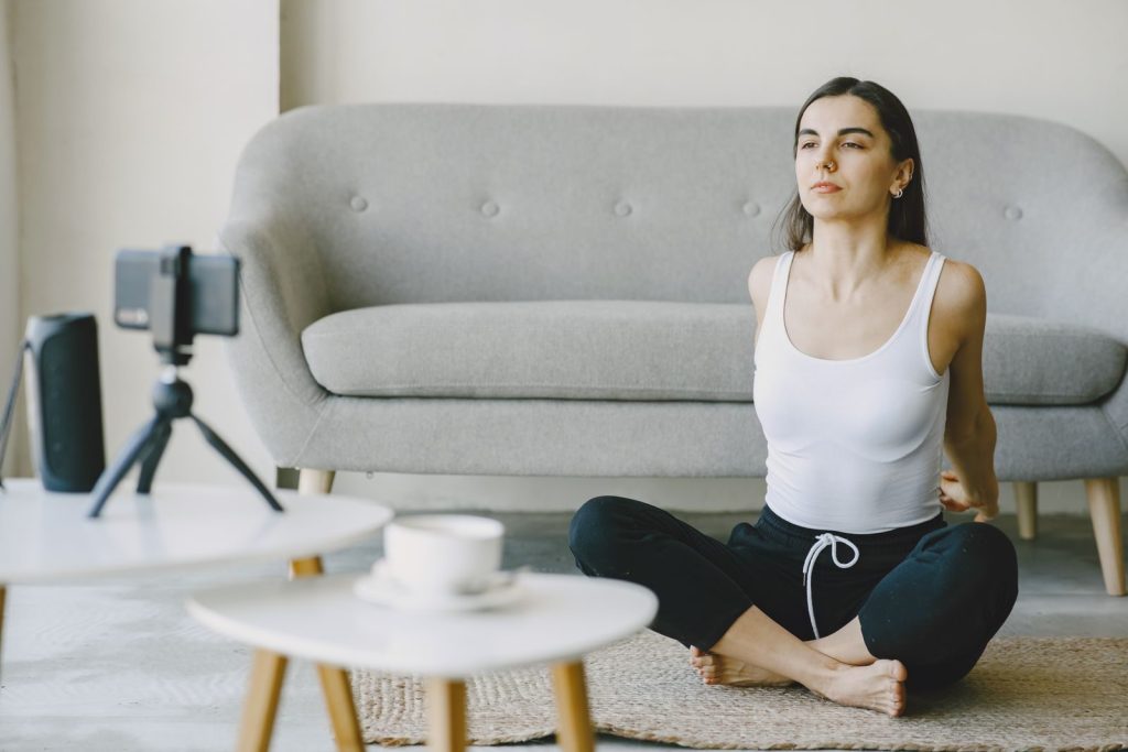 Young woman practicing yoga at home with a smartphone for virtual guidance, sitting cross-legged in a serene living room.