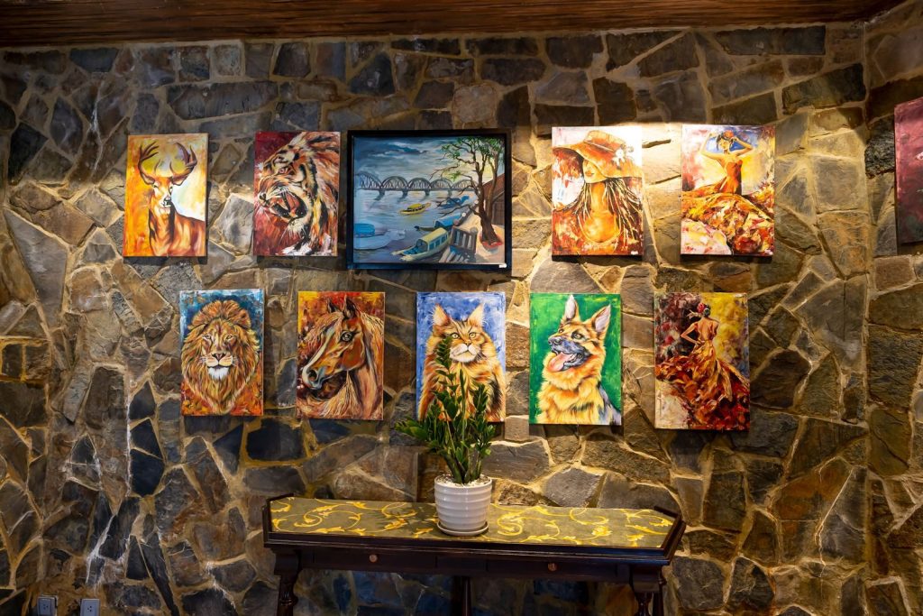 A vivid indoor display of animal and landscape paintings on a textured stone wall.