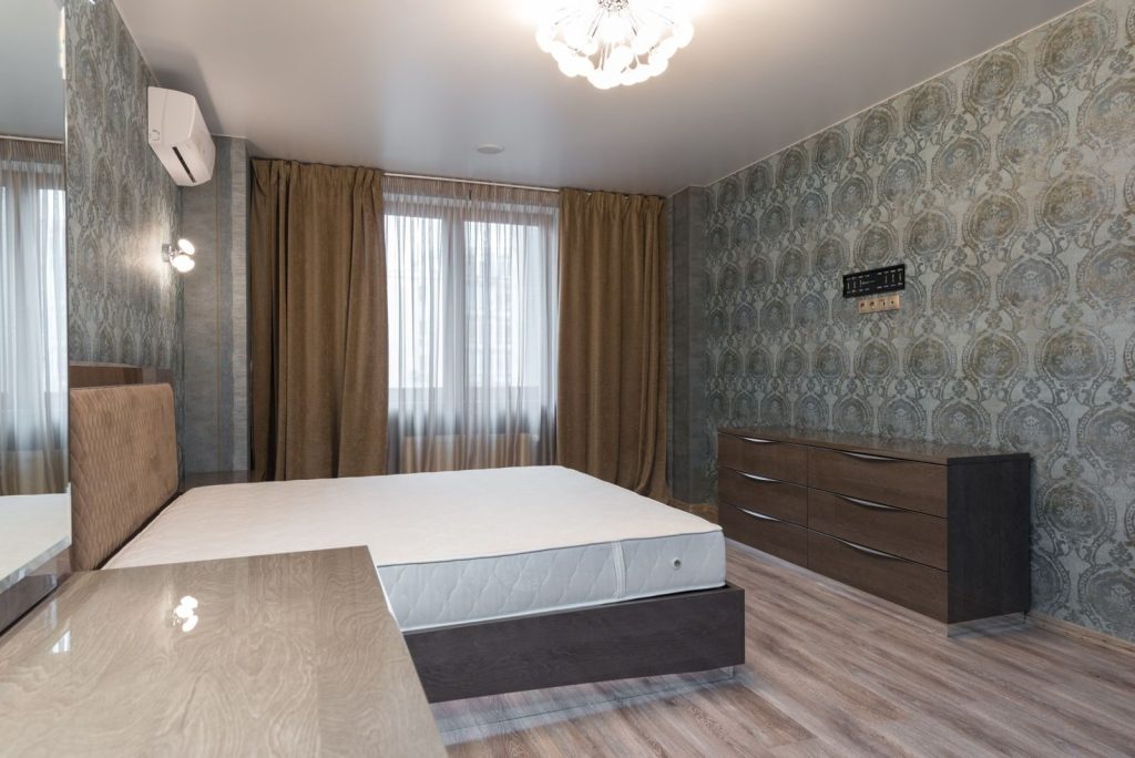 Interior of bedroom with air conditioning above bed placed near window with curtains and dresser