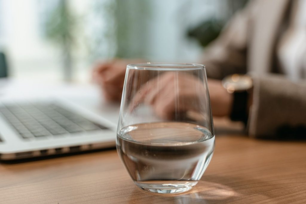 Close-up of a glass of water on a desk in an office setting with a laptop in the background.