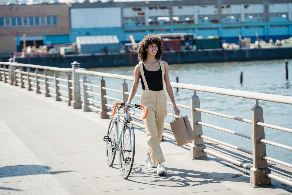 Full body of content black woman with bicycle and shopping bag walking near fence on embankment in daylight