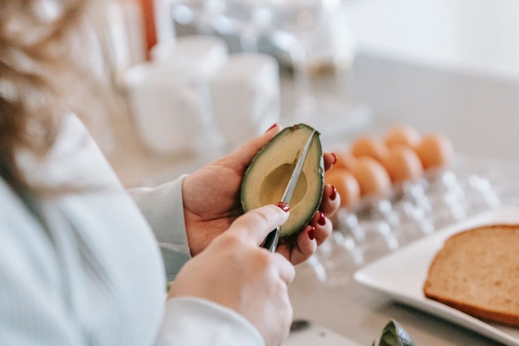 Crop unrecognizable female with knife cutting fresh avocado while making toasts at table with eggs in light kitchen during morning