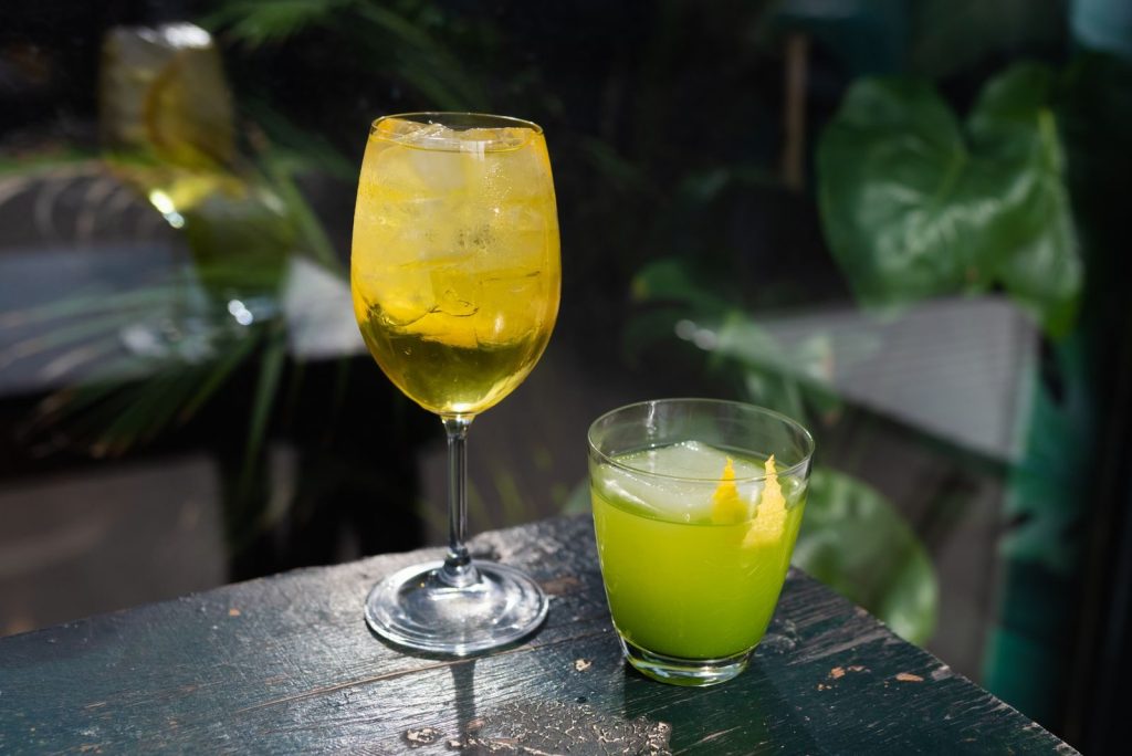 Two vibrant cocktails on a wooden table with ice and lemon garnishes, set in natural light.