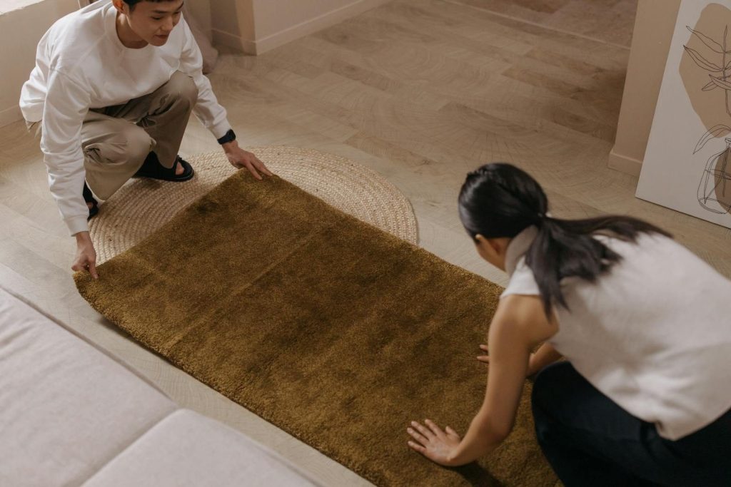 A man and woman collaboratively arranging a brown rug on a wooden floor inside a home.