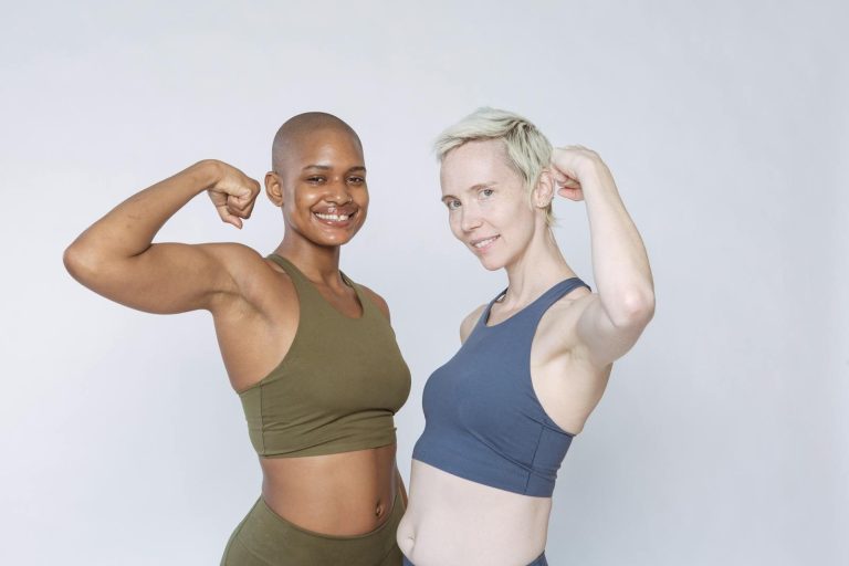 Cheerful diverse fit females in sportswear pumping biceps muscle and looking at camera while standing against white background
