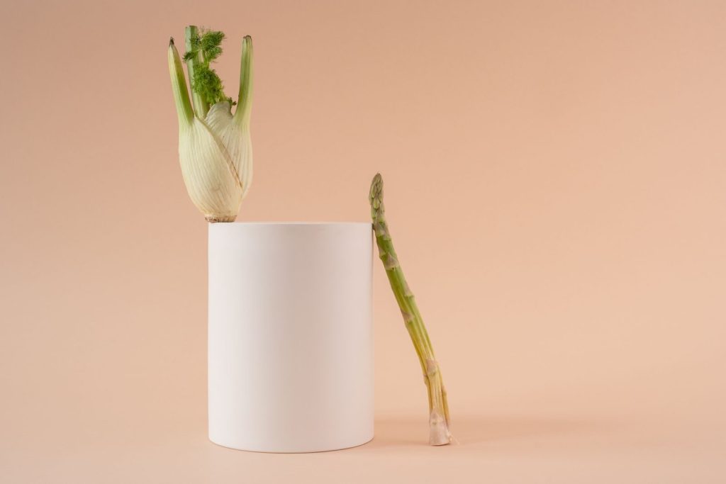 A minimalist still life image featuring asparagus and fennel on a geometric white cylinder against a beige background.