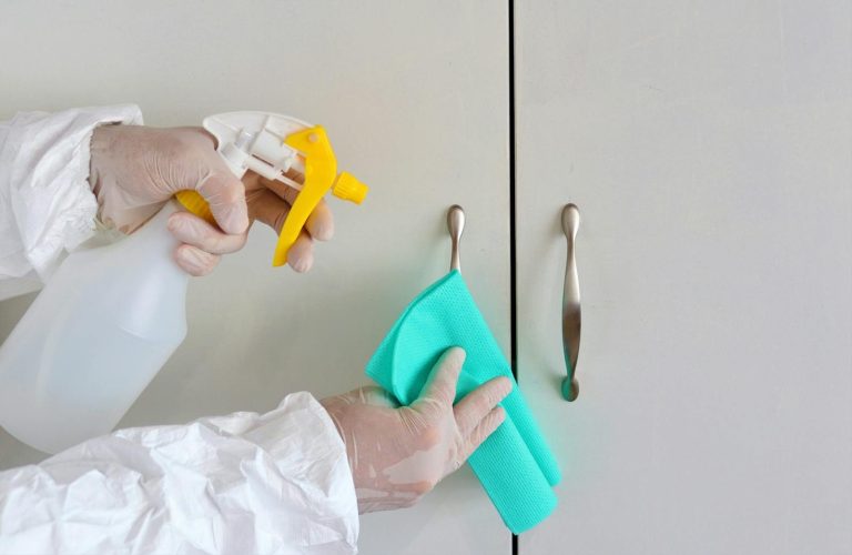 Gloved hands cleaning cabinet handles with a spray bottle and cloth for hygiene.
