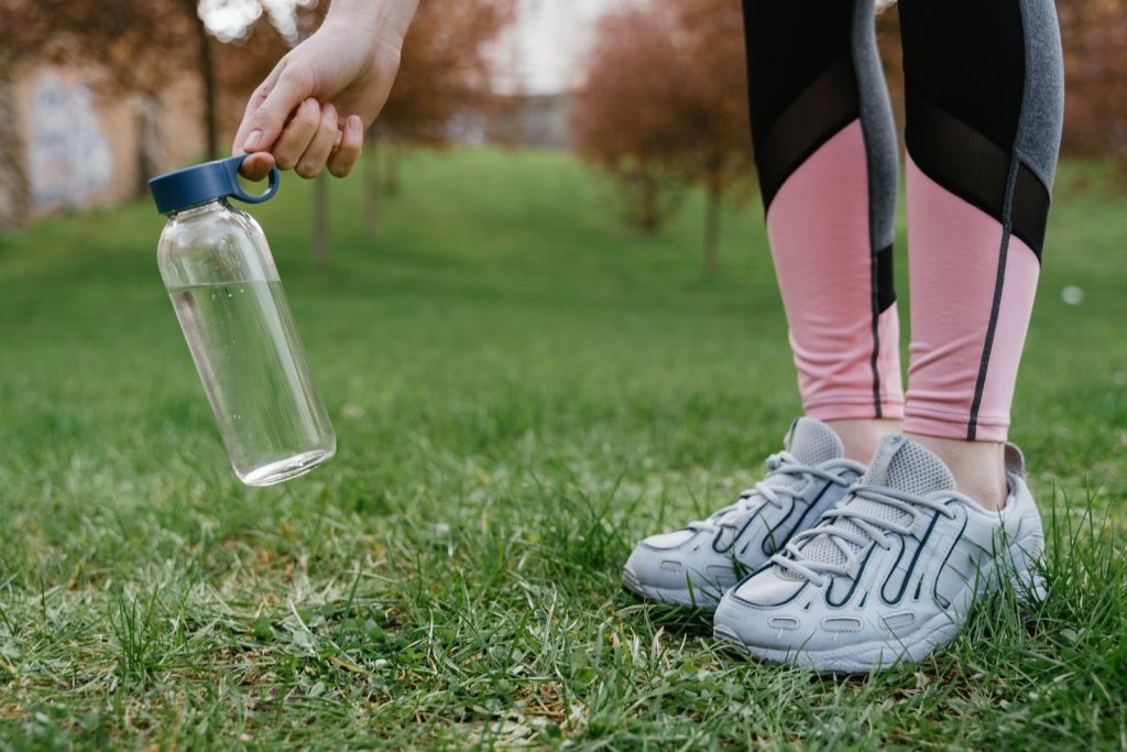 Close-up of legs in pink leggings and sneakers with a glass water bottle in hand on grass.