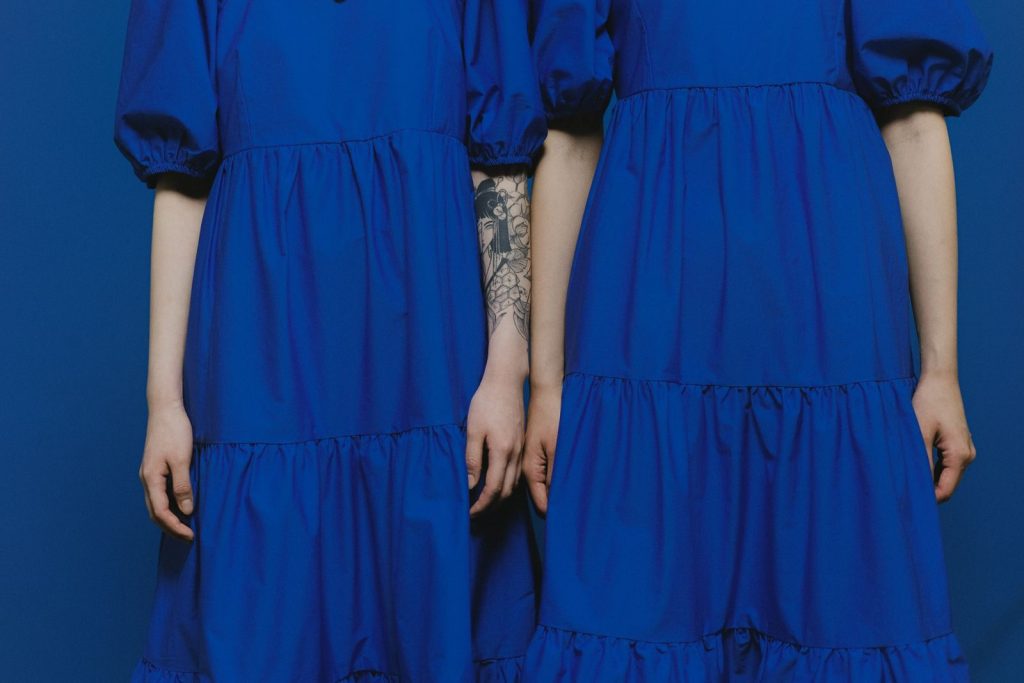 Close-up of two women standing side by side wearing blue dresses, one with a tattoo.