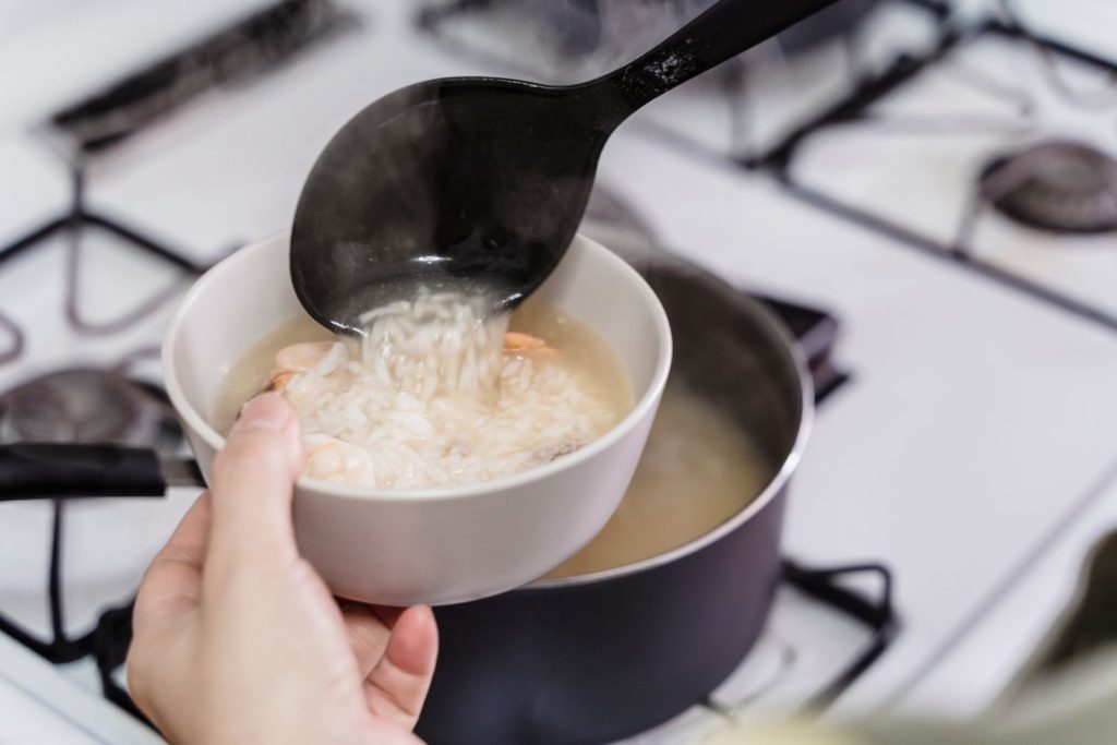 A person ladling steaming rice soup into a bowl over a stove, capturing a moment of home cooking.