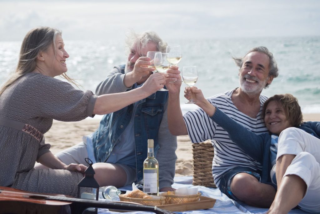 Group of senior friends enjoying a beach picnic in Portugal, toasting wine glasses and enjoying laughter.